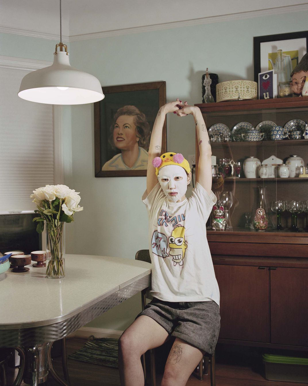 A tattooed woman, a beauty mask on her face, arms stretched above her head.