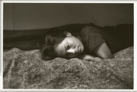 A black and white portrait of a woman lying face down on a bed, gazing off to the side.