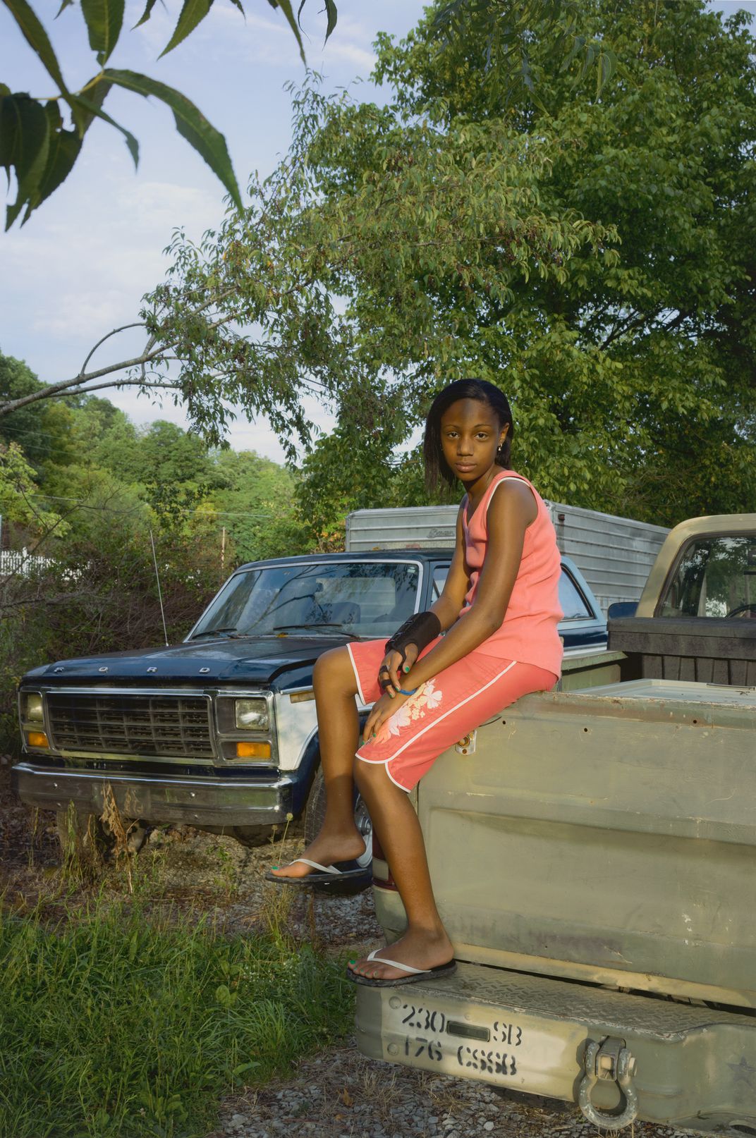 A portait of a black girl with a wrist brace wearing a pink tanktop and shorts while sitting on a the edge of a truck bed.